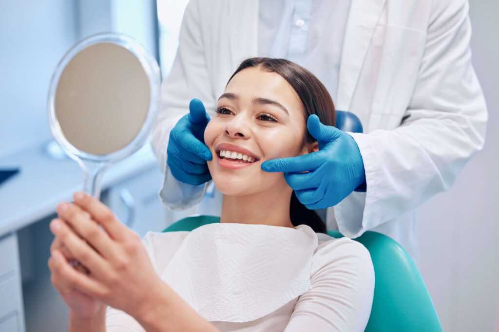 Dentist in blue gloves touching corners of woman's smile as she looks into mirror